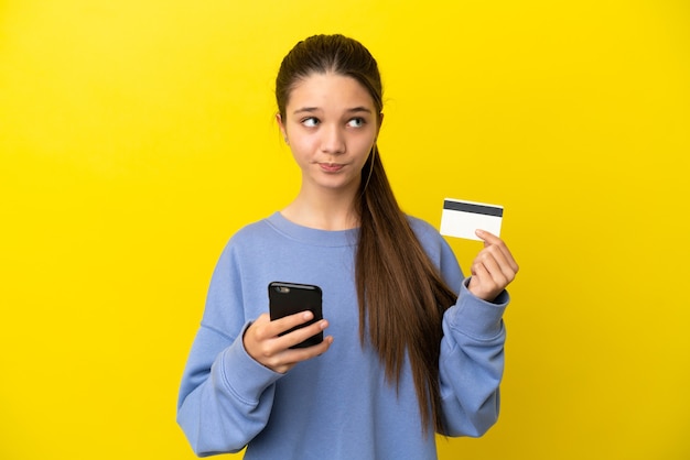 Niña sobre fondo amarillo aislado comprando con el móvil con tarjeta de crédito mientras piensa