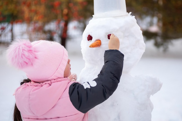 Niña sola haciendo cara de muñeco de nieve con medios improvisados durante el día mientras camina en el parque con árboles y luz solar brillante en el fondo Los padres pasan tiempo con los niños