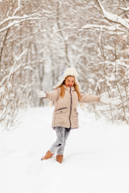 Niña con sombrero blanco y chaqueta en el bosque de invierno arroja nieve. Diversión de invierno