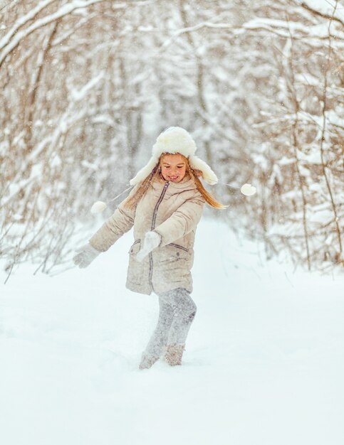 Niña con sombrero blanco y chaqueta en el bosque de invierno arroja nieve. Diversión de invierno