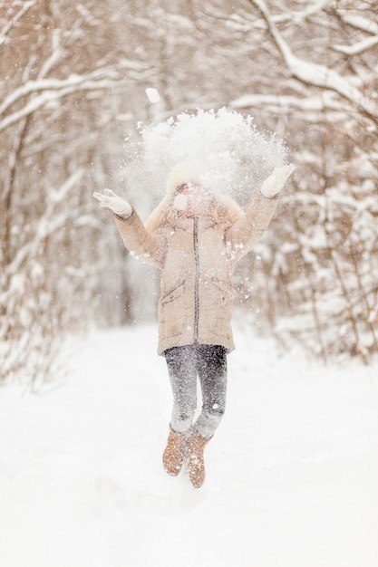 Niña con sombrero blanco y chaqueta en el bosque de invierno arroja nieve. Diversión de invierno