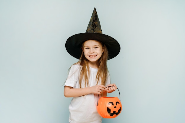 Una niña con sombrero de bruja sostiene una canasta de dulces sobre un fondo azul y sonríe Truco o trato