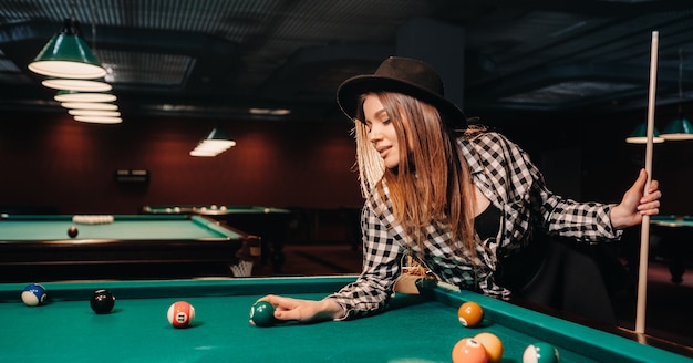 Una niña con un sombrero en un club de billar con un taco y bolas en sus manos. Jugar al billar.