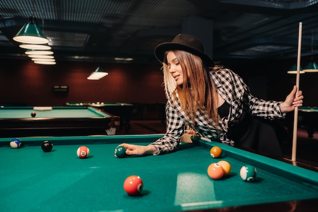 Una niña con un sombrero en un club de billar con un taco y bolas en sus manos.