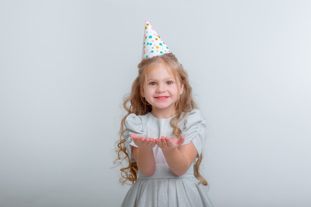 Niña con sombrero de cumpleaños soplando confeti en blanco