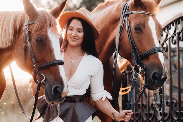 Una niña con un sombrero se encuentra junto a los caballos en la naturaleza.