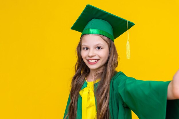 Una niña con un sombrero de graduado muestra un selfie de un graduado Un niño brillante con el traje de un maestro Fondo amarillo aislado