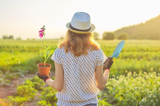 Niña con sombrero con guantes caminando en el jardín con pala y planta de flor en maceta