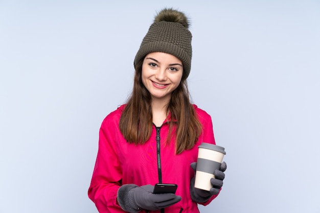 Niña con sombrero de invierno aislado en la pared azul con café para llevar y un móvil