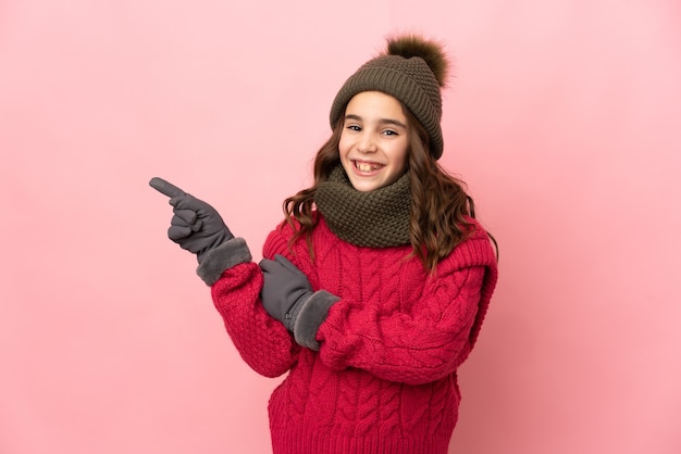 Niña con sombrero de invierno aislado en la pared rosada apuntando con el dedo hacia el lado