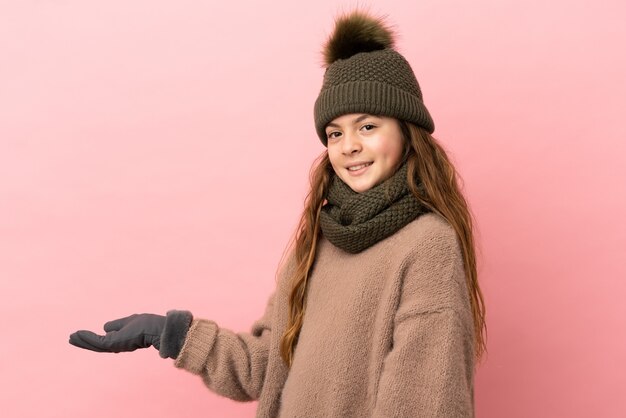 Niña con sombrero de invierno aislado sobre fondo rosa extendiendo las manos hacia el lado para invitar a venir