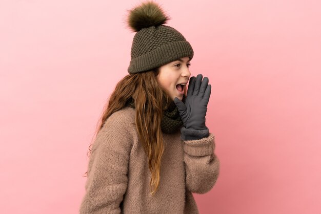 Niña con sombrero de invierno aislado sobre fondo rosa gritando con la boca abierta hacia el lado