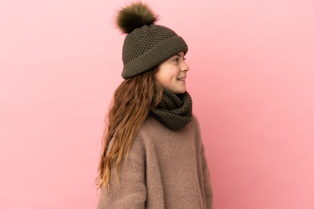 Niña con sombrero de invierno aislado sobre fondo rosa mirando de lado