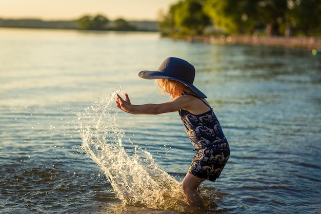 niña en un sombrero junto al río en verano salpica agua