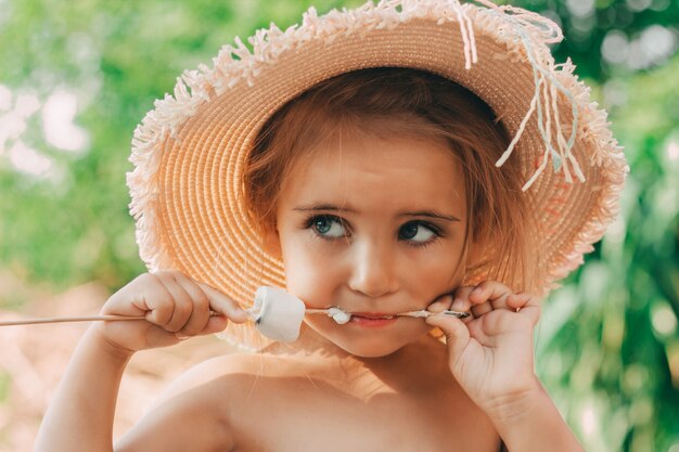 Niña con sombrero de paja comiendo malvaviscos aireados malvaviscos fritos sobre el fuego