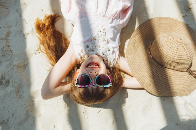 Una niña con un sombrero de paja y gafas de sol está tomando el sol en una playa de arena