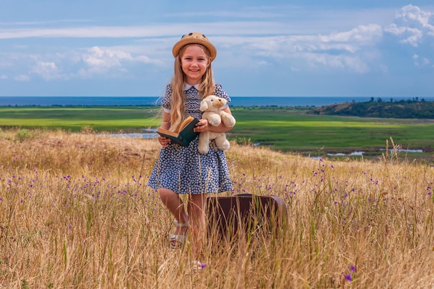 Niña con sombrero de paja y vestido sentado en una maleta vintage y leyendo un libroNiño lindo con juguete mirando el cuaderno