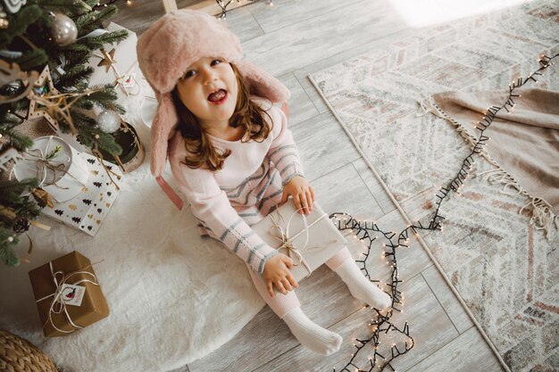 Niña con un sombrero de piel de invierno se sienta en el suelo cerca del árbol de Navidad con regalos.