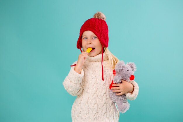 niña con un sombrero de punto rojo, guantes y un suéter blanco sonríe en una pared azul