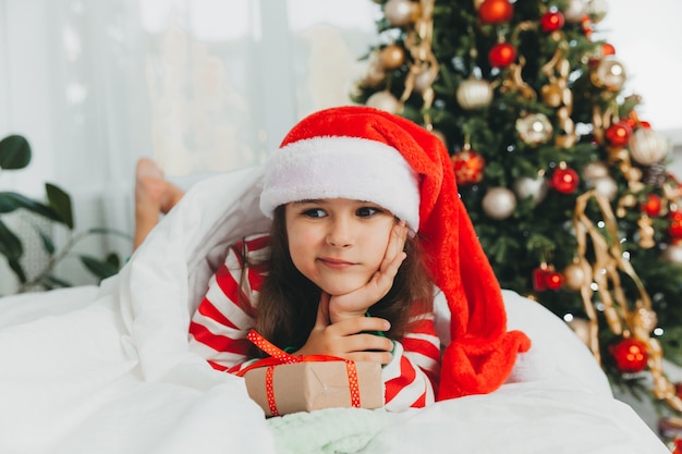 Niña con sombrero rojo de año nuevo con un regalo de Navidad. Ella se acuesta en la cama, abrazada a una caja contra el fondo de un árbol de Navidad.