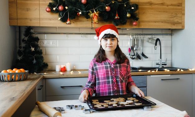 Una niña con un sombrero rojo y una camisa a cuadros roja está de pie en la cocina junto a una bandeja de galletas