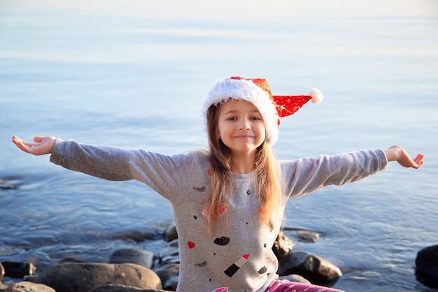 Una niña con un sombrero rojo de Navidad se sienta en la orilla del mar con los brazos extendidos. Viajar por vacaciones de invierno