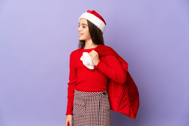 Niña con sombrero y saco de Navidad aislado en la pared púrpura mirando hacia el lado y sonriendo