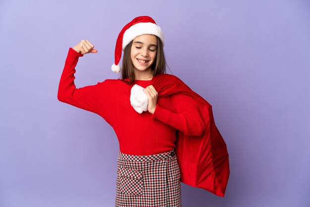 Niña con sombrero y saco de Navidad aislado sobre fondo púrpura haciendo gesto fuerte