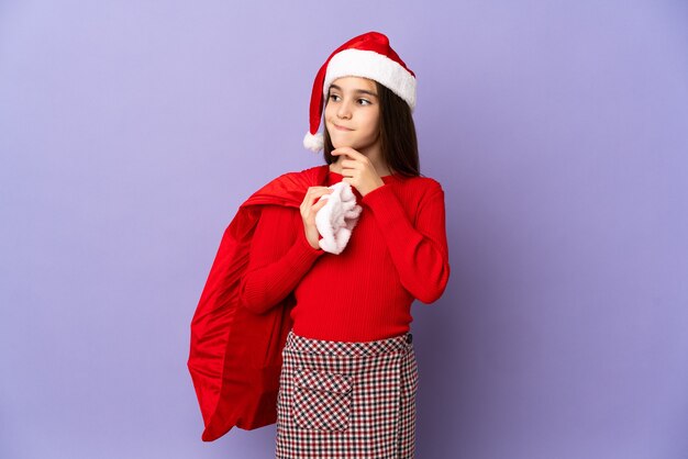 Niña con sombrero y saco de Navidad aislado sobre fondo púrpura teniendo dudas y pensando