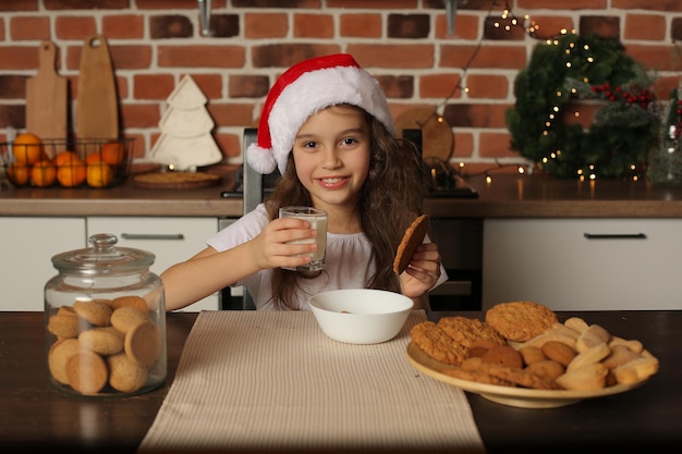 Una niña con un sombrero de Santa Claus en la cocina tiene galletas y un vaso de leche en sus manos