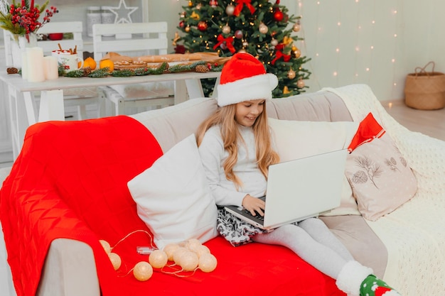 Una niña con un sombrero de santa claus se comunica por videollamada a través de una computadora portátil, sentada en el sofá de la sala de estar.