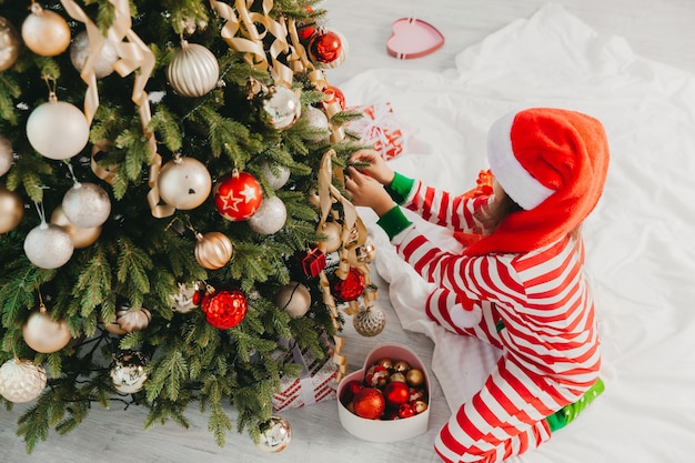 Niña con sombrero de santa claus decora el árbol con juguetes. Un lindo bebé se prepara para celebrar la Navidad.