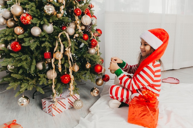 Niña con sombrero de santa claus decora el árbol con juguetes. Un lindo bebé se prepara para celebrar la Navidad.