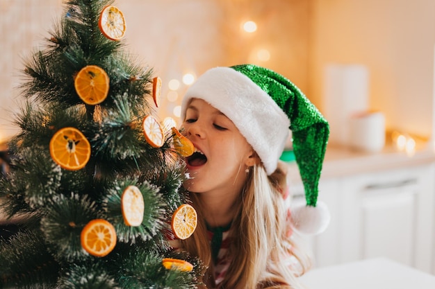 Una niña con un sombrero de santa claus decora el árbol con mitades secas de naranja. Un lindo bebé se prepara para celebrar la Navidad.