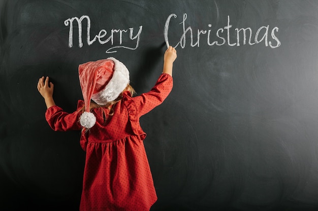 Una niña con sombrero de santa claus dibuja dibujos navideños en la pizarra