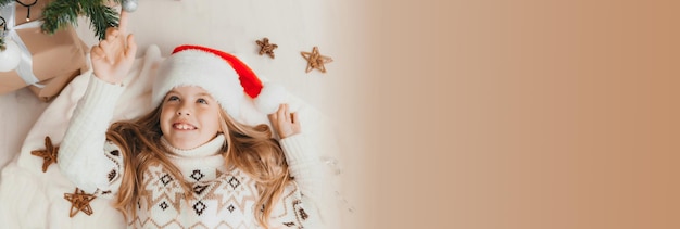 Niña en un sombrero de santa claus yace cerca del árbol de navidad en casa vista desde arriba Navidad