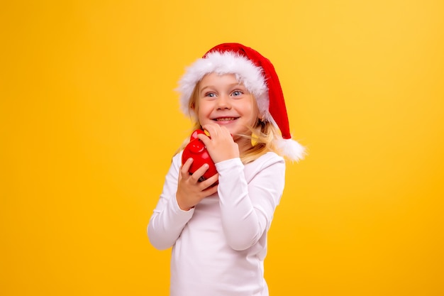 Niña con sombrero de Santa con hucha roja en la pared amarilla, espacio para texto