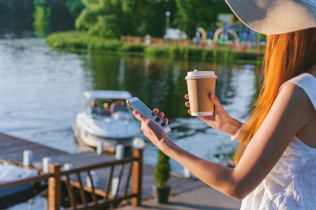 Una niña con sombrero sostiene un teléfono inteligente con café en sus manos. En los terraplenes de agua de fondo con una embarcación de recreo.