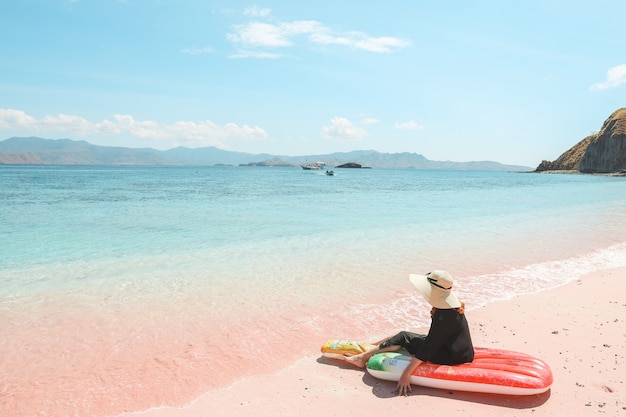 Una niña con sombrero de verano sentado en un colchón inflable mientras disfruta de la vista al mar en la playa de arena rosa