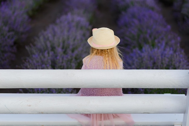 Una niña con sombrero y vestido rosa está sentada en un banco en un campo de lavanda