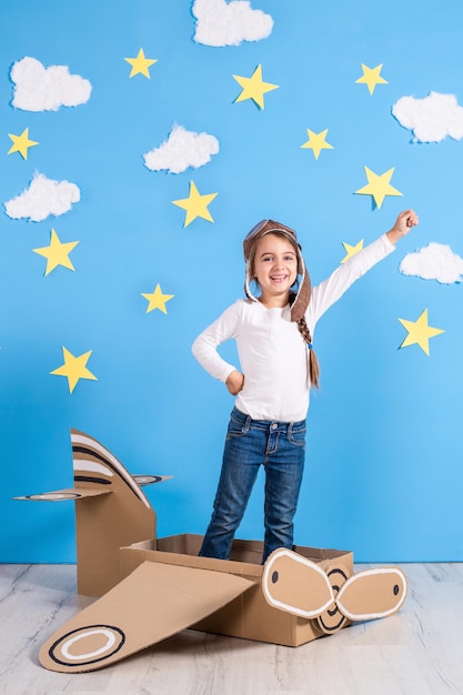 Niña soñadora jugando con un avión de cartón en el estudio con fondo de cielo azul y nubes blancas.