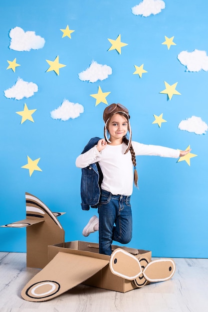 Niña soñadora jugando con un avión de cartón en el estudio con fondo de cielo azul y nubes blancas.