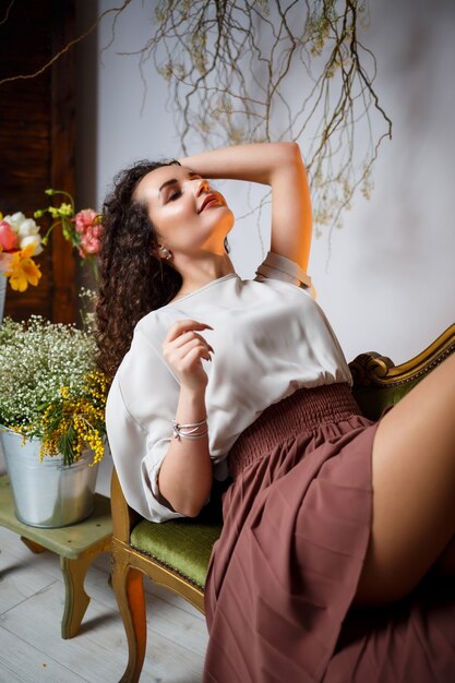 La niña sonríe con una falda marrón sentada en el sofá. alrededor hay flores frescas en un jarrón.