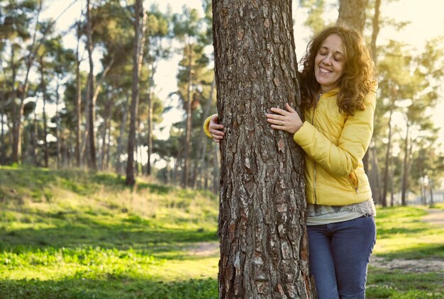 Niña sonriendo con los ojos cerrados con chaqueta amarilla y pantalón azul desde el frente abrazando un árbol en un bosque de pinos al atardecer