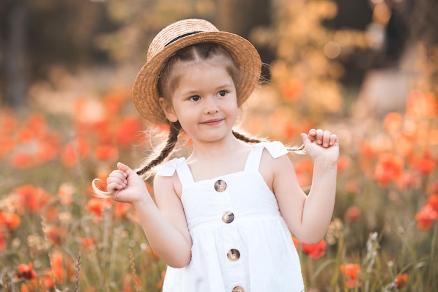 Una niña sonriente de 3 a 4 años usa sombrero de paja y vestido rústico blanco posando sobre un fondo de amapola natural. Niño feliz en el prado de flores. tiempo de primavera Infancia.