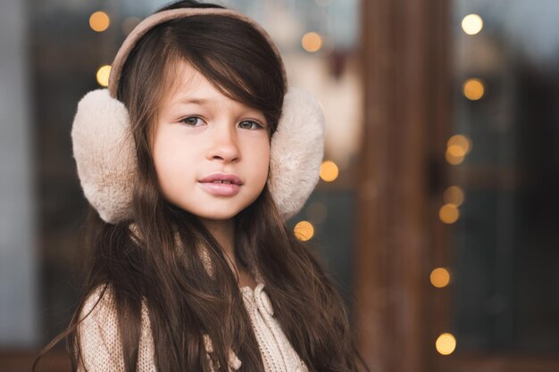 Niña sonriente de 4-5 años con auriculares esponjosos sobre luces brillantes de Navidad de cerca. Mirando a la cámara. Infancia. Año nuevo. Temporada de vacaciones de invierno.