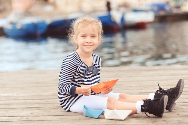 Niña sonriente de 5 a 6 años sosteniendo un barco de papel en el fondo del mar. Mirando a la cámara. Infancia.