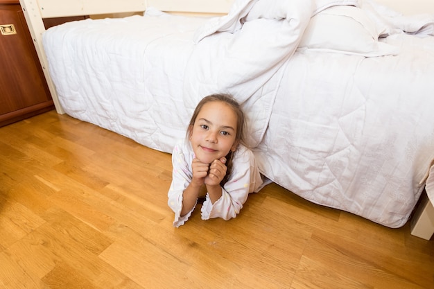 Niña sonriente acostada debajo de la cama en casa y mirando a la cámara