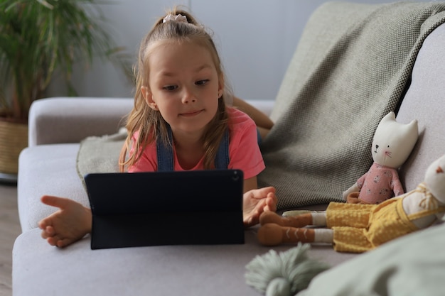 Niña sonriente acostada en el sofá jugando juegos en línea, información de navegación web, usando aplicaciones divertidas en tableta.