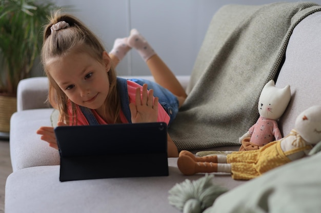 Niña sonriente acostada en el sofá jugando juegos en línea, información de navegación web, usando aplicaciones divertidas en tableta.